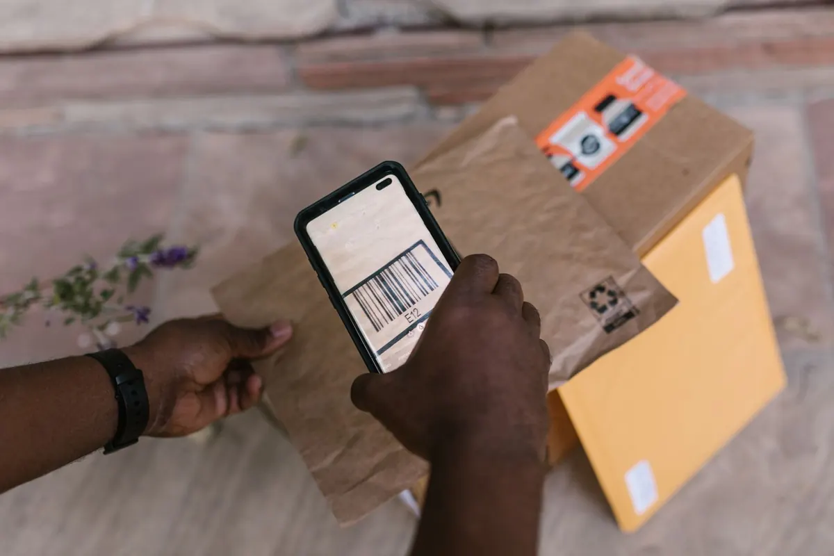 A hand holding a smartphone that scans the barcode on a brown paper parcel lying on a yellow padded envelope