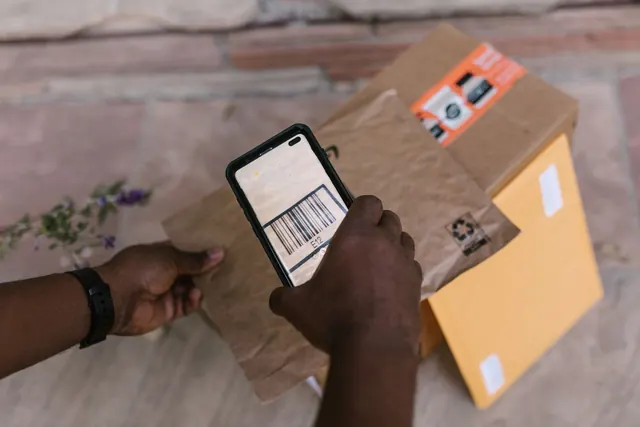 A hand holding a smartphone that scans the barcode on a brown paper parcel lying on a yellow padded envelope