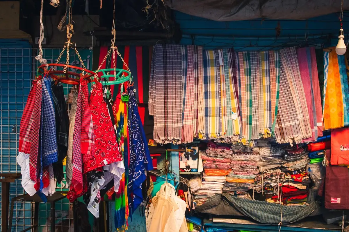 A market stall hung with checked and striped cotton cloths, red and blue printed scarves and piles of folded fabric under a bare bulb