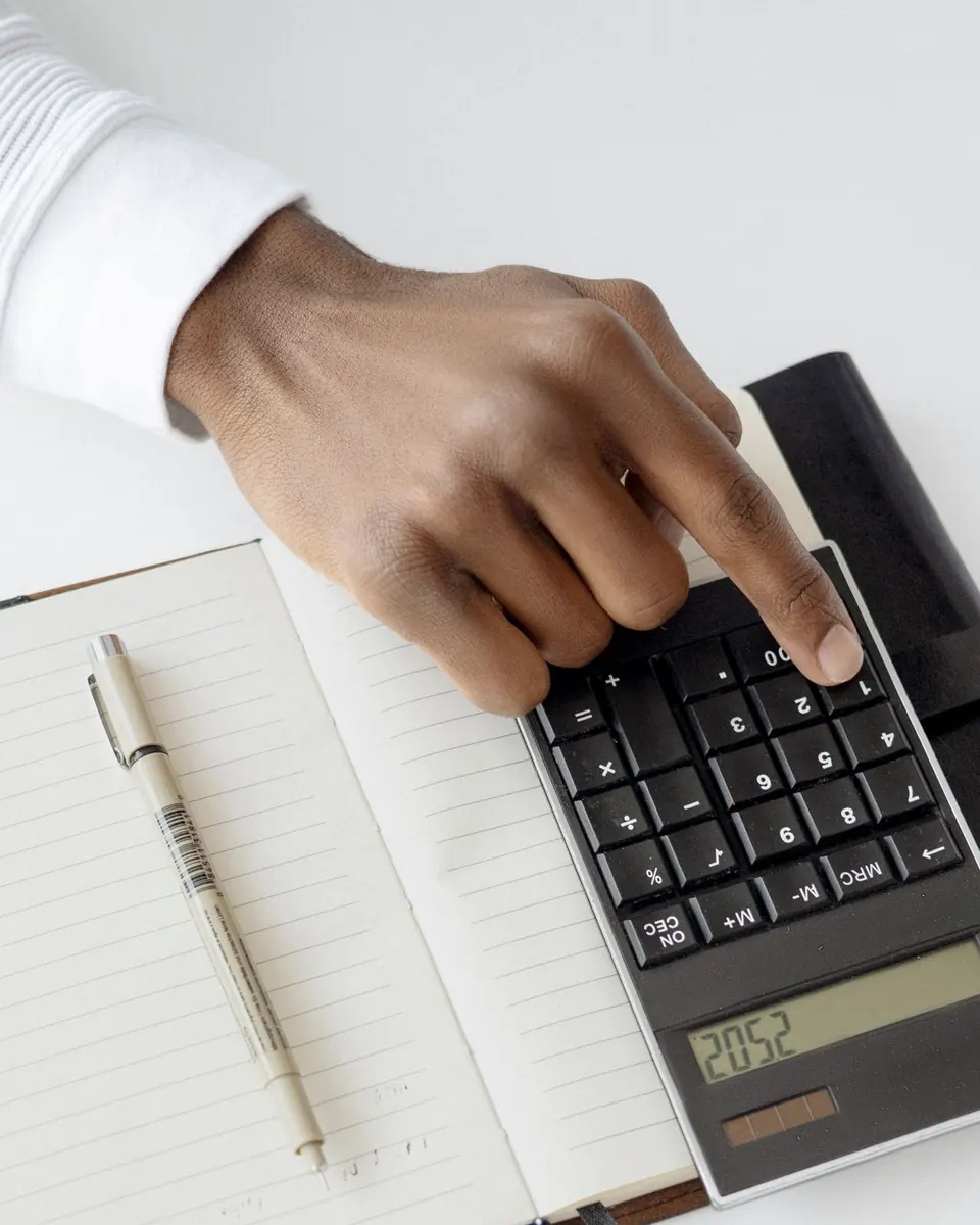 A finger pressing a key on a black desk calculator that sits on an open lined notebook with a pen beside it