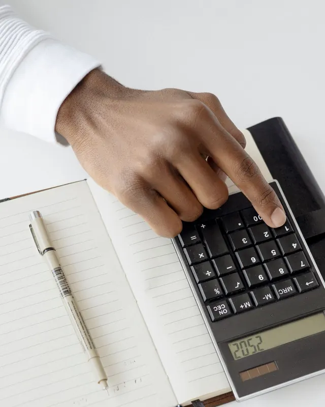 A finger pressing a key on a black desk calculator that sits on an open lined notebook with a pen beside it