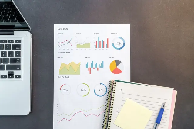 A printed page of line, bar, area and pie charts on a dark desk next to a laptop keyboard, a spiral notebook, a sticky note and a blue pen