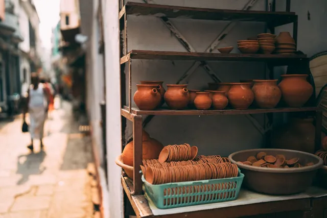 Terracotta pots, small clay cups and lamps on metal shelves of a shop stall beside a narrow alley, with a blurred person walking away.