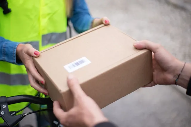 One person in a yellow high-visibility vest passes a small cardboard box with a barcode sticker to another person; only their hands and arms are in the frame