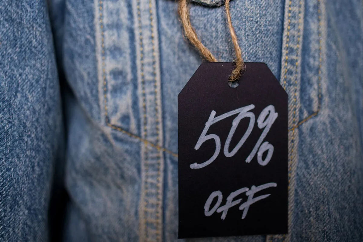 A black paper tag on twine hangs on a denim jacket with 50% OFF handwritten in white pen.