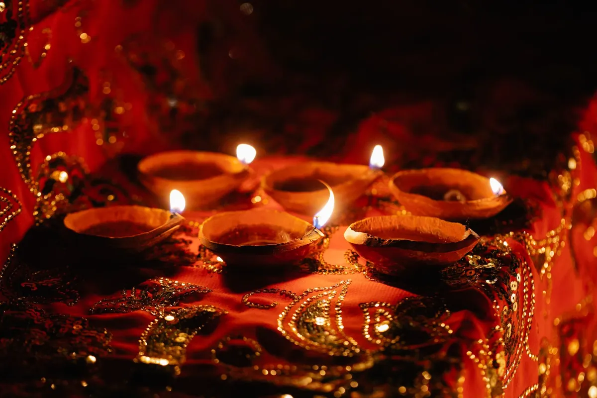 Six lit clay diyas stand on red fabric embroidered with gold sequins in a dark room.