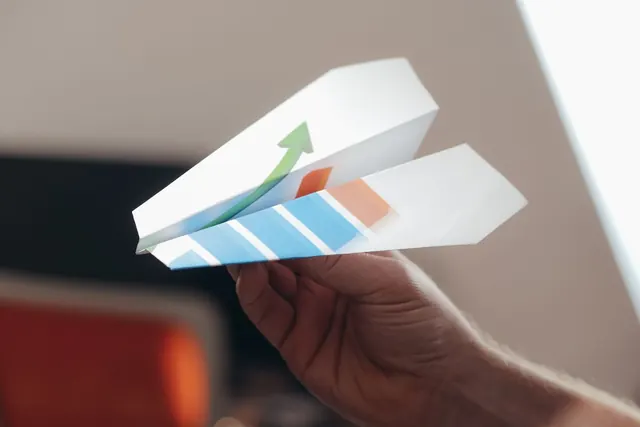 A hand holding a white paper plane printed with blue and orange bars and a green upward arrow
