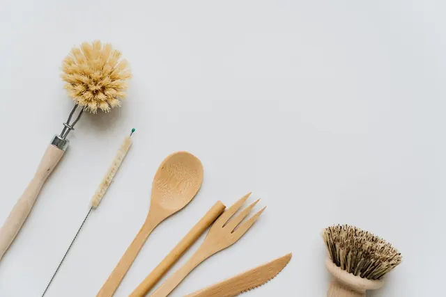 A flat lay of a wooden spoon, fork and knife with two dish brushes and a thin cleaning brush on a pale grey surface