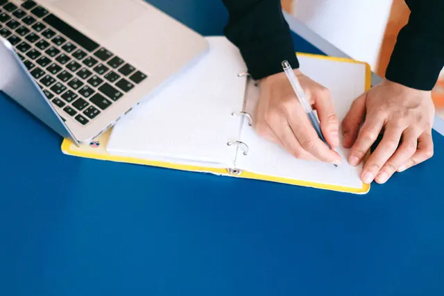 Hands writing with a pen in a yellow ring binder next to an open silver laptop on a blue desk