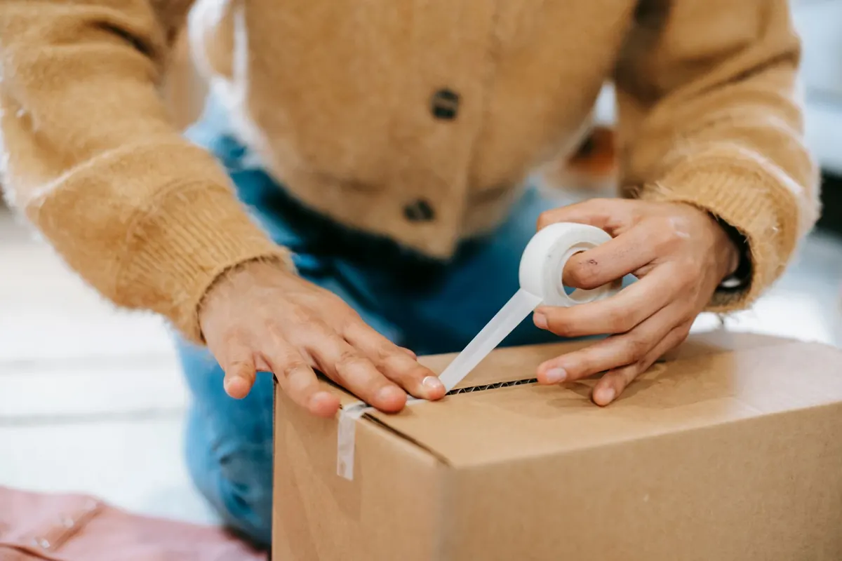 Hands pulling clear tape across the top of a brown cardboard box while a person in a mustard cardigan kneels behind it