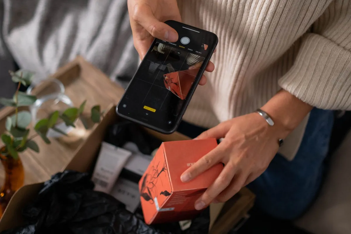 Hands hold a smartphone above an open cardboard box to photograph a small orange product box, with only the sleeve and torso of the person in view.