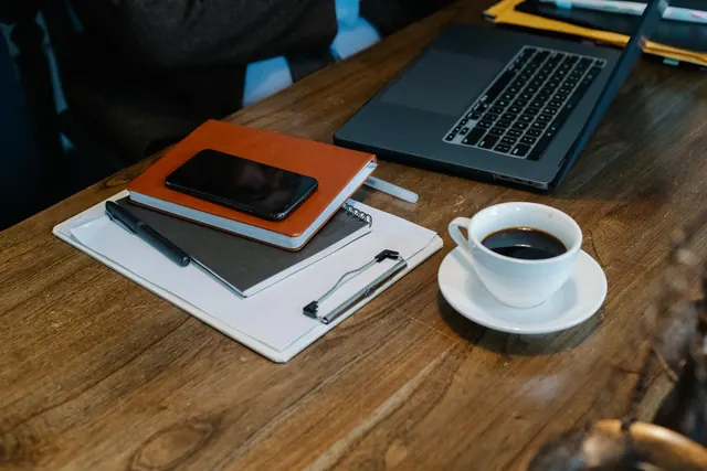 A white cup of black coffee on a saucer beside a clipboard, notebooks and a smartphone on a worn wooden table with a laptop behind