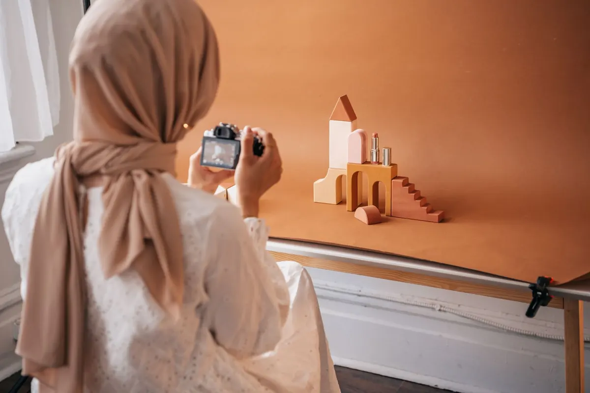 A person in a beige headscarf, seen from behind, photographing small wooden blocks and a lipstick set on an orange paper backdrop