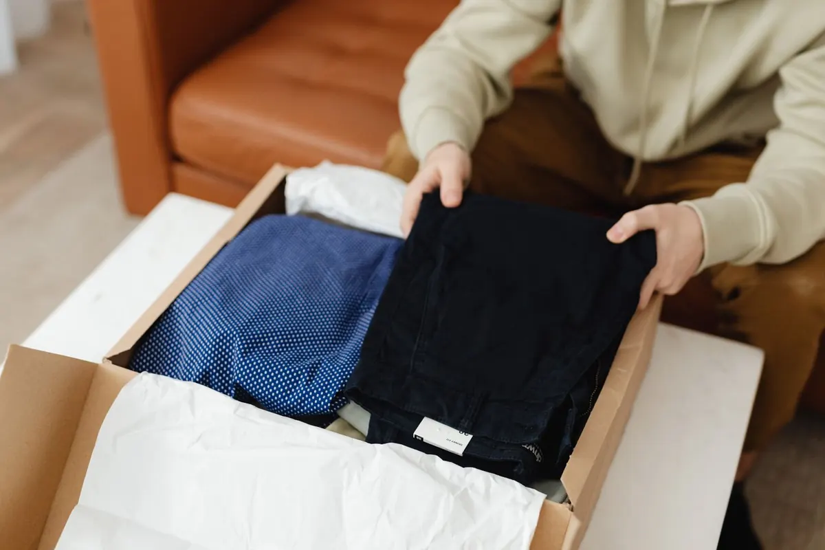 An open cardboard box holding a folded blue spotted shirt and dark trousers on white tissue paper, with a person's hands lifting the trousers