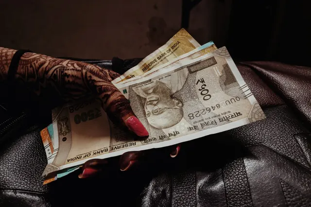 A hand decorated with henna and red nail polish holding a fan of Indian five-hundred-rupee and other rupee notes over a dark leather bag