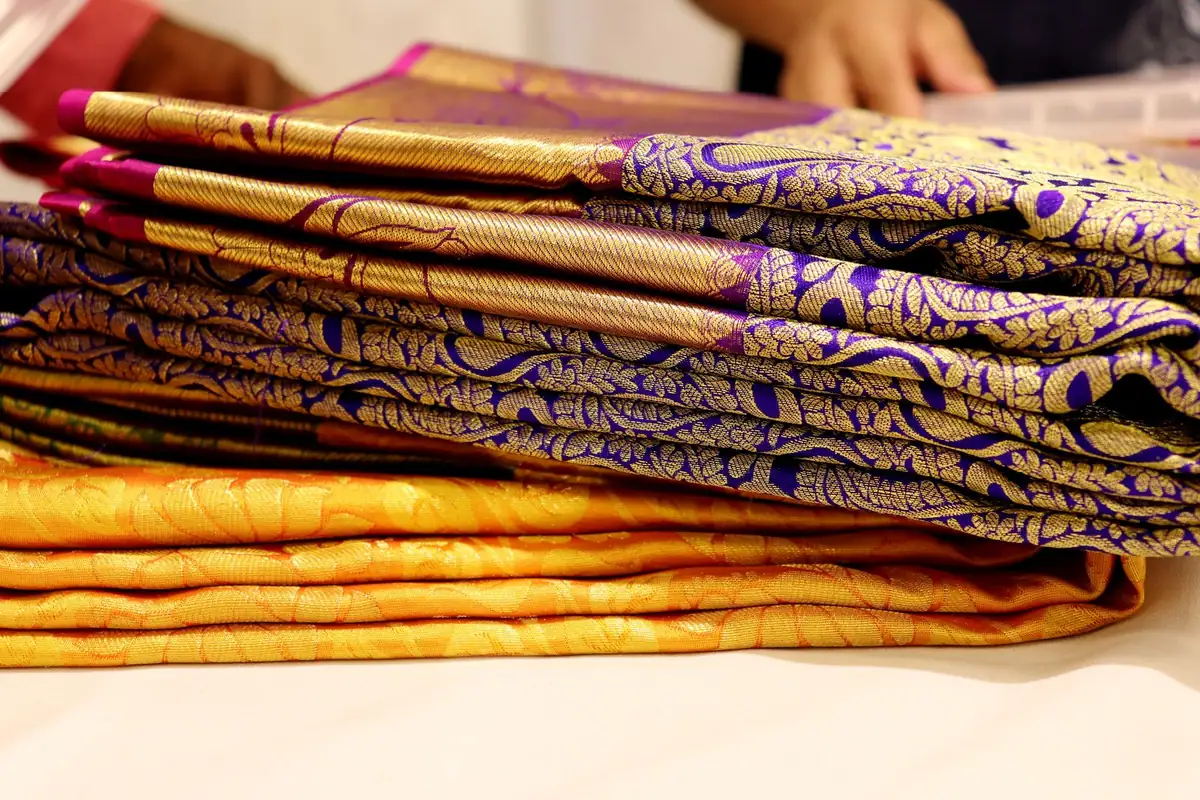 A pile of folded silk sarees with purple and gold patterned borders lying on top of an orange saree