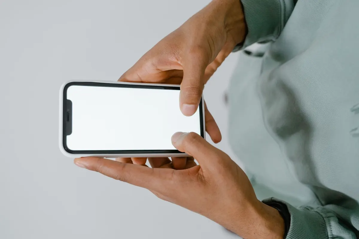 A person in a sage-green sweatshirt holding a smartphone sideways with both hands, its screen blank white